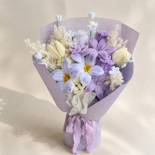 Lavender, pale blue and ivory handmade chenille flower bouquet with lilac wrapping and trailing ribbons on a warm ivory background.