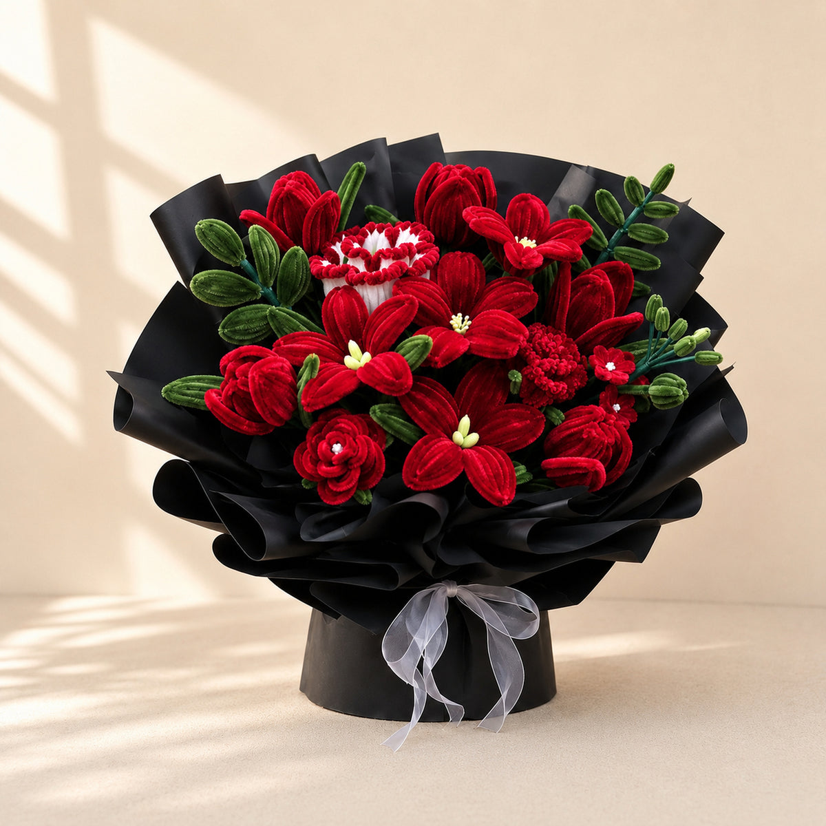Deep red and dark green handmade chenille flower bouquet with black wrapping and a translucent white ribbon on a warm neutral background.
