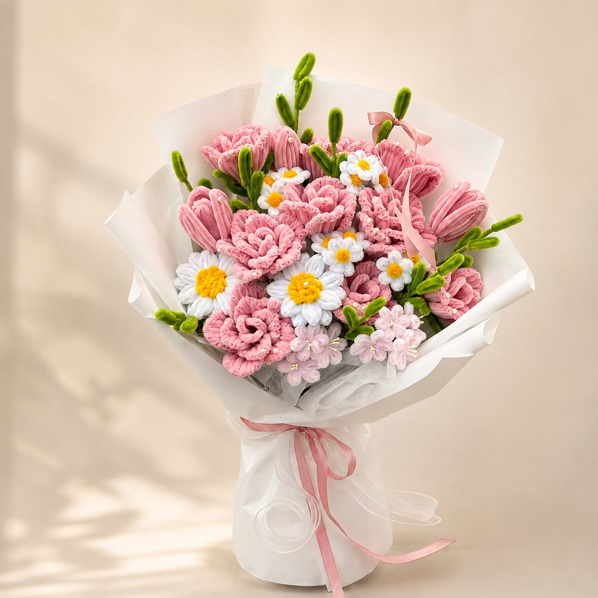 Pink and white handmade chenille flower bouquet with daisy-style blooms, green buds, white wrapping and pink ribbons on a warm neutral background.